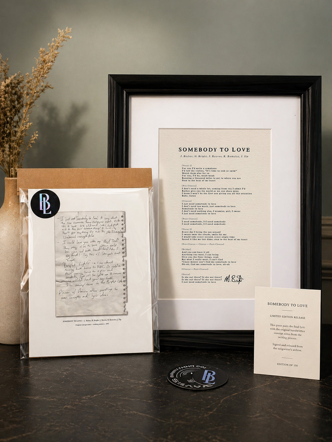 Framed document with 'Somebody to Love' text and a vase with dried flowers on a dark surface.